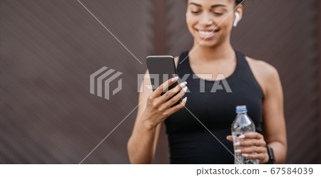 African american girl with headphones and fitness tracker holds bottle of water and smartphone African american girl with headphones and fitness tracker holds bottle of water and smartphone 67584039
