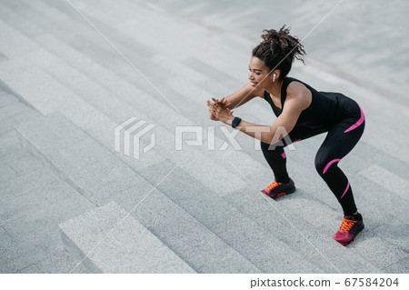 Girl with wireless headphones and fitness tracker doing exercises on stairs in city 67584204