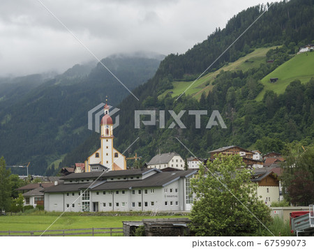 Summer morning foggy view on Neustift im Stubaital church at Stubaital or Stubai Valley near Innsbruck, Austria, green meadow and misty mountain peaks 67599073