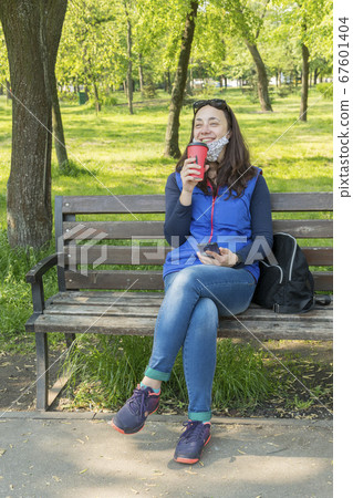 The woman sitting on the bench in the park and wearing a protective mask and using smartphone . Coronavirus concept. toned. vertical photo The woman sitting on the bench in the park and wearing a protective mask and using smartphone . Coronavirus concept. toned. vertical photo 67601404