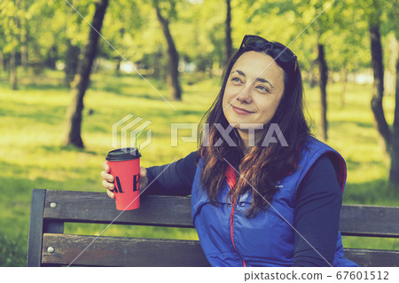 Beautiful Caucasian woman with long brown hair sitting in the park drinking a cup of tea or coffee and smiling. Middle-aged athletic woman with long hair drinks coffee in a park. toned 67601512
