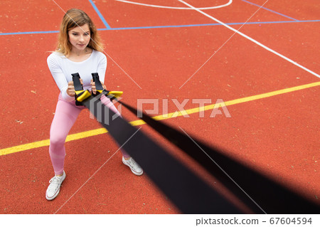 Pretty young Caucasian girl in pink sportswear exercising with suspension trainer sling, doing squats, outdoors at red sport court. Fitness concept with trx trainer 67604594