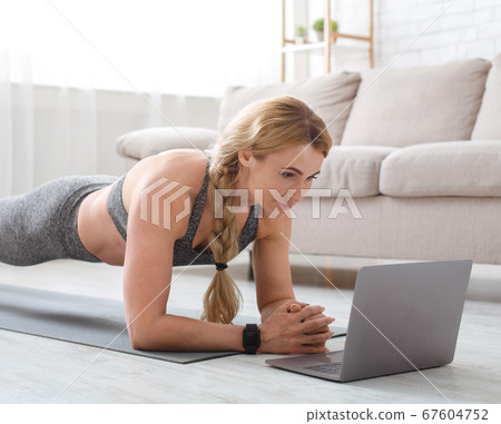 Middle aged woman in sportswear with fitness tracker makes plank at mat on floor and looks at laptop 67604752