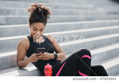 African american girl in sportswear with fitness tracker looks in smartphone, near bottle of water 67604884