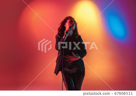 Young caucasian musician, female singer performing on gradient background in neon light. Concept of music, hobby, festival Young caucasian musician, female singer performing on gradient background in neon light. Concept of music, hobby, festival 67606511