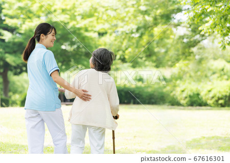 Rear view of senior woman and caregiver rehabilitating outdoors Rear view of senior woman and caregiver rehabilitating outdoors 67616301