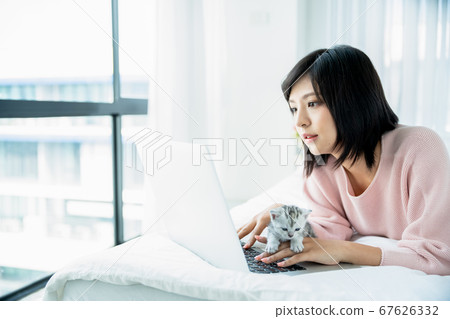 Cute woman uses a computer while playing with a kitten in a bright bedroom 67626332