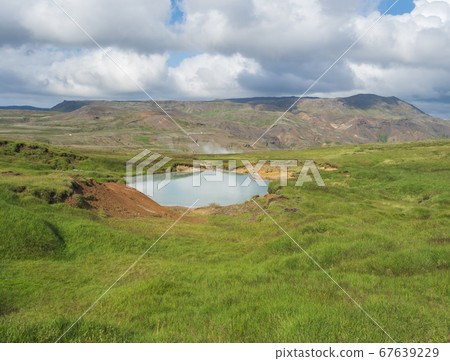 Reykjadalur valley with hot springs river and pool with lush green grass meadow and hills with geothermal steam. South Iceland near Hveragerdi city. Summer sunny morning, blue sky. 67639229