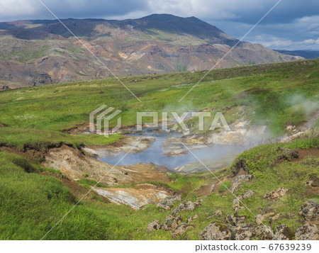 Reykjadalur valley with hot springs river and pool with lush green grass meadow and hills with geothermal steam. South Iceland near Hveragerdi city. Summer sunny morning, blue sky. 67639239