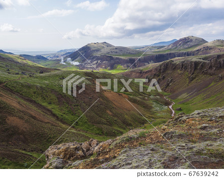 Reykjadalur valley with hot springs river with lush green grass meadow and hills with geothermal steam. South Iceland near Hveragerdi city. Summer sunny morning, blue sky. 67639242