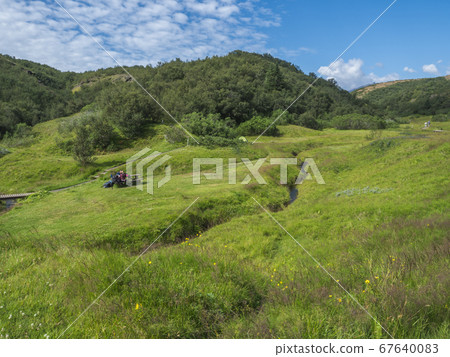 view on lush green grass of Langidalur camping site in Thorsmork with hikers sitting on wooden pinic table bench. Highlands of Iceland, end of the Laugavegur hiking trail. Summer Blue sky. view on lush green grass of Langidalur camping site in Thorsmork with hikers sitting on wooden pinic table bench. Highlands of Iceland, end of the Laugavegur hiking trail. Summer Blue sky. 67640083
