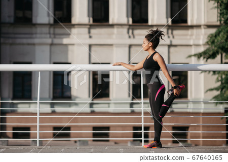Athlete woman preparing for run on city street. African american girl in sportswear doing warm up of leg muscles in stadium in morning Athlete woman preparing for run on city street. African american girl in sportswear doing warm up of leg muscles in stadium in morning 67640165