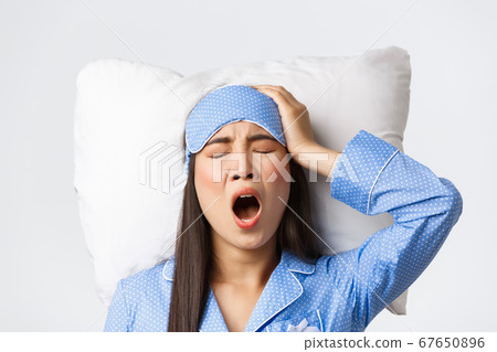 Close-up of tired and sleepy cute asian girl in sleeping mask and pajamas, yawning and touching head as feeling headache after hangover sleepover party, having insomnia problems, lying on pillow 67650896
