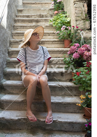 Beautiful female tourist wearing big straw sun hat and shorts sitting and relaxing on old stone house stairs during summer travel on Mediterranean cost on hot summer day Beautiful female tourist wearing big straw sun hat and shorts sitting and relaxing on old stone house stairs during summer travel on Mediterranean cost on hot summer day 67652998