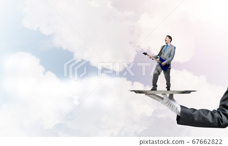 Businessman on metal tray playing electric guitar against blue sky background Businessman on metal tray playing electric guitar against blue sky background 67662822
