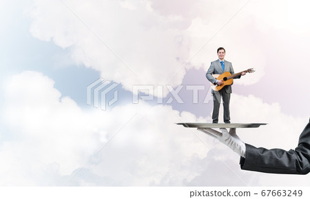 Businessman on metal tray playing acoustic guitar against blue sky background Businessman on metal tray playing acoustic guitar against blue sky background 67663249