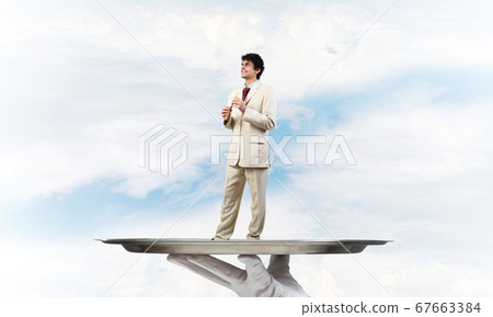 Businessman on metal tray playing fife against blue sky background Businessman on metal tray playing fife against blue sky background 67663384