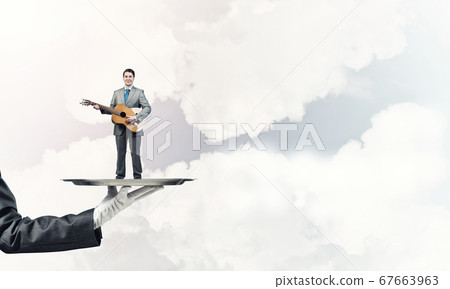 Businessman on metal tray playing acoustic guitar against blue sky background Businessman on metal tray playing acoustic guitar against blue sky background 67663963