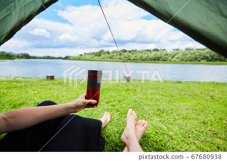 Two pairs of legs stick out from tent on a grassy glade meadow and near river. Two pairs of legs stick out from tent on a grassy glade meadow and near river. 67680938