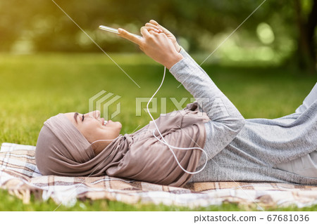 Cheerful muslim girl resting at park, watching videos on smartphone Cheerful muslim girl resting at park, watching videos on smartphone 67681036