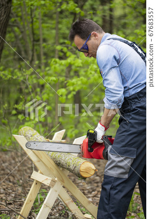 Woodcutter in working uniform sawing tree trunk on sawhorse with electric saw Woodcutter in working uniform sawing tree trunk on sawhorse with electric saw 67683557