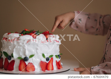 Little girl first time taste bithday cake. Cute little girl in beautiful dress tasting cake with strawberries. Close up of girl put finger into cake 67684470