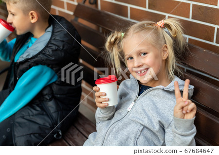 Two cute adorable little caucasian blond children enjoy sitting on wooden bench and drink hot chocolate drink with paper cup. Beatiful happy smiling 7 years old child portrait 67684647