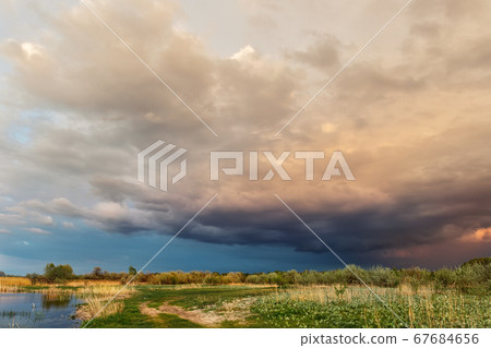 Dramatic dark cloudy thunder sky over dry grass field at pond shore. Rural countryside rainy ominous cloudscape over dry autumn grass field. Dirt country road landscape with bad weather 67684656