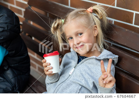 Cute adorable little caucasian blond kid girl enjoy sitting om wooden bench and drink hot chocolate drink with paper cup. Beatiful happy smiling 7 years old child portrait Cute adorable little caucasian blond kid girl enjoy sitting om wooden bench and drink hot chocolate drink with paper cup. Beatiful happy smiling 7 years old child portrait 67684667