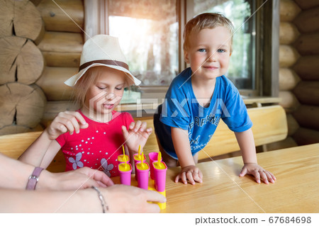 Two cute adorable caucasian blond little sibling sitting at table home yard and have fun tasting mom homemade fruit icecream. Young happy sister and brother enjoy laughing outside. Family love care 67684698