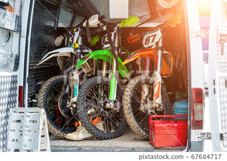 Three bright dirty sport motocross motorcycles stand in tail of technical support assistance van in team camp during extreme offroad championship. Mobile cycle service, maintenance transport concept 67684717