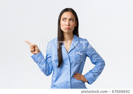 Pouting disappointed asian girl in blue pajamas, frowning and sulking as pointing looking upper left corner dissatisfied, stare with dislike, complaining as standing white background 67688692