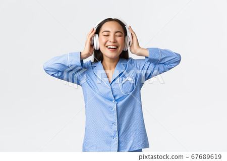 Home leisure, weekends and lifestyle concept. Happy pleased asian woman in pajamas enjoying awesome sound quality in new earphones, dancing in pajamas and listening music 67689619
