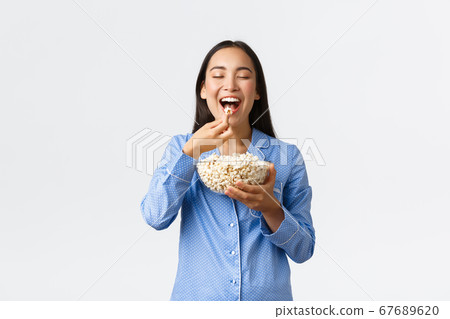 Home leisure, sleepover and slumber party concept. Smiling delighted asian girl enjoying eating favorite popcorn, wearing pajama on weekend, watching favorite movie, white background Home leisure, sleepover and slumber party concept. Smiling delighted asian girl enjoying eating favorite popcorn, wearing pajama on weekend, watching favorite movie, white background 67689620