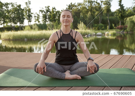 Enjoying life. Young beautiful and happy woman in sportswear and wireless earphones sitting in lotus pose on a mat and meditating in the morning near lake in park 67690605