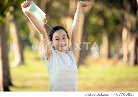 Happy smiling asian child girl is standing with raised arms up,holding mask in her hand in green nature,woman enjoy breathing fresh air after Covid-19 quarantine,Coronavirus ended,end of stay home 67700855