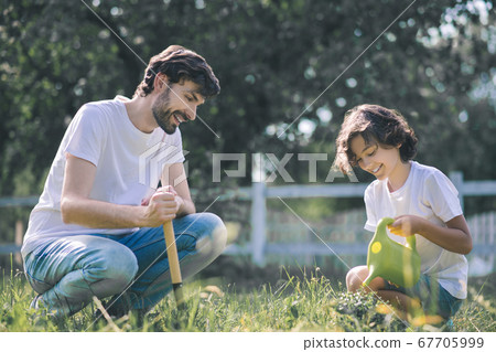Dark-haired boy watering the plant and smiling, his father watching him 67705999