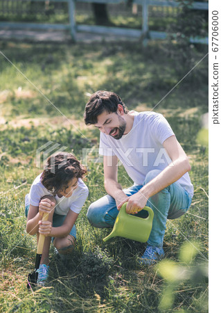 Dark-haired man watering the plant, his son watching him Dark-haired man watering the plant, his son watching him 67706000
