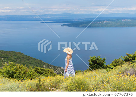 Rear view of young woman wearing striped summer dress and straw hat standing in super bloom of wildflowers, relaxing while enjoing beautiful view of Adriatic sea nature, Croatia 67707376