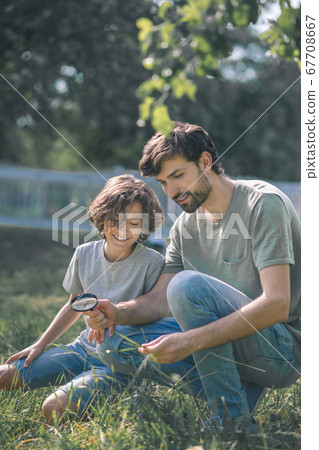 Dark-haired boy and his father looking at the plant through magnifying glass and looking interested Dark-haired boy and his father looking at the plant through magnifying glass and looking interested 67708667