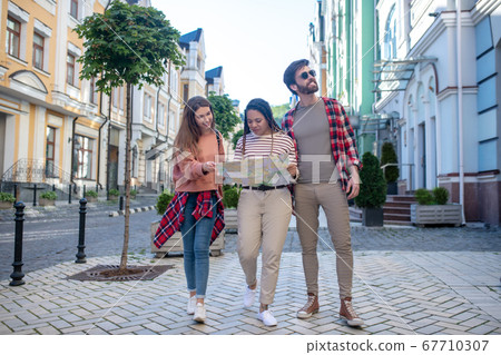 Two girls with map and guy in sneakers walking along street Two girls with map and guy in sneakers walking along street 67710307