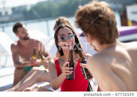 Beautiful dark-haired woman in red swimming suit drinking beer and talking to a man Beautiful dark-haired woman in red swimming suit drinking beer and talking to a man 67710329