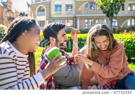 Laughing guy and two girls sitting on plaid on the grass Laughing guy and two girls sitting on plaid on the grass 67710343