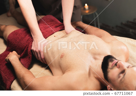 Male masseur massages the thoracic diaphragm of a muscular male athlete in a massage room with dark lighting 67711067