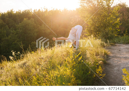 Young yoga woman trainer standing Forward Bend with Shoulder Opener in evening at sunset. 67714142