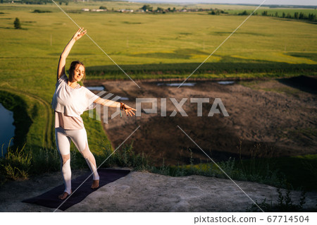 Caucasian woman practicing yoga on top of rock background of flowing river and green meadow 67714504