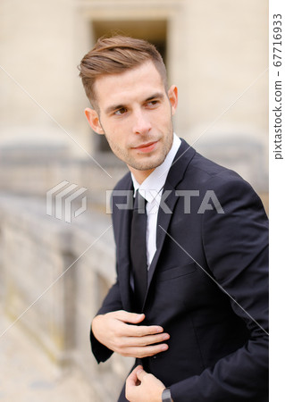 Portrait of young man wearing black suit and leaning on banister. 67716933