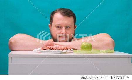 Attractive fat young man in casual t-shirt makes easy choice between green apple or sausage 67725139
