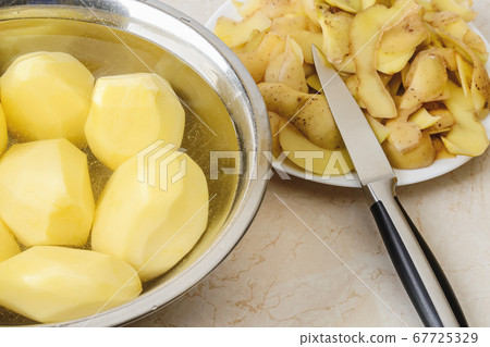 Raw peeled potatoes in a bowl with water, potato peels and a vegetable knife on a plate. 67725329