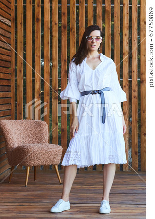 Young stylish sexy woman in summer white dress standing on wooden terrace in tropical hotel, palm trees background, long black hair, sunglasses, earrings, sunglasses 67750869